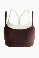 Top deportivo en tejido funcional de color marrón con tirantes dobles en color crudo y diseño de superposición. Cuenta con escote redondo y copas con relleno extraíble.