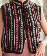 Chaleco tejido a mano en crochet de lana merino color negro, con diseño sin mangas, cuello mao y cierre delantero con cordones para atar. Presenta un patrón de rayas verticales con detalles geométricos en tonos fucsia, celeste y blanco.