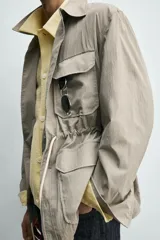 Parka relaxed fit color gris, confeccionada en tejido técnico ligero, con cuello solapa, manga larga con puño abotonado, cuatro bolsillos de plastrón con solapa en el frente, cintura ajustable con cordón y cierre frontal de botones.