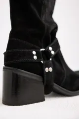 Bota de caña media confeccionada en cuero gamuzado color chocolate. Presenta un diseño de estilo western con taco cuadrado de 7 cm y una correa decorativa en el tobillo con herrajes metálicos plateados.