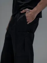 Pantalón cargo negro de corte oversize, confeccionado en microfibra elastizada. Presenta cintura elástica, bolsillos laterales y un bolsillo cargo con cierre de broche a presión. Incluye un sistema de elásticos con tancas en los tobillos para ajustar la apertura.