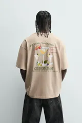 Camiseta de manga corta y cuello redondo, color beige claro, con corte relajado (relaxed fit) y un pequeño texto bordado en el pecho.