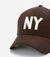 Gorra de seis paneles con ala curva, confeccionada en algodón color marrón. Presenta un bordado frontal con las letras NY en color blanco y cuenta con cierre strapback ajustable en la parte posterior.