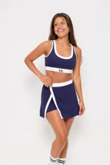 Conjunto deportivo compuesto por un top y una pollera short. El top es de escote redondo con ribetes blancos y banda elástica inferior con logo 'bk.'. La pollera short presenta un diseño cruzado con ribetes blancos y cintura elástica.