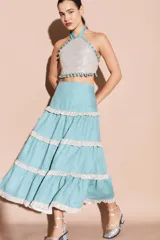 Falda larga de lino color turquesa con volados horizontales decorados con ribetes brillantes en tono plateado. Presenta una silueta amplia y fluida.