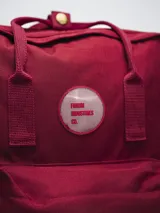 Mochila color verde militar con diseño clásico y minimalista. Cuenta con un compartimento principal con cierre de doble cremallera, un bolsillo frontal con cierre y dos asas superiores con broche. En el frente, tiene un parche circular con el texto "FORUM INDUSTRIES CO." en rojo.