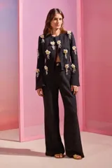 Chaqueta de punto jacquard color negro con hilos de lúrex, corte entallado, cuello redondo y mangas ligeramente acampanadas. Presenta flores blancas y doradas bordadas a mano en toda la superficie y cierre frontal con gancho en el cuello.