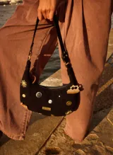 Cartera baguette de gamuza negra adornada con diversos dijes y tachas metálicas en forma de sol, botones y animales, con correa de hombro y placa con logo metálico.