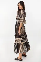 Vestido largo de algodón con patrón de rayas verticales y bloques de colores en tonos marrón, negro y crudo. Presenta diseño cruzado tipo envolvente con solapas negras, mangas tres cuartos acampanadas con volados, falda escalonada y lazo negro ajustable en la cintura.