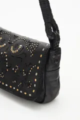 Cartera de mano tipo baguette confeccionada en cuero vacuno negro, con solapa decorada mediante diseño calado y tachas metálicas. Presenta una correa trenzada y un pañuelo estampado anudado en uno de sus laterales.