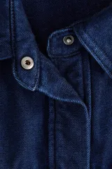 Camisa de denim rígido de algodón azul oscuro, con corte relajado, hombros caídos, cuello clásico, botones ocultos a presión, bolsillos superiores con solapa y bajo redondeado.