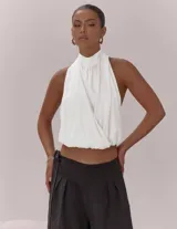Top blanco con diseño halter, cuello alto y espalda descubierta. Presenta un frente cruzado con pliegues, bajo elástico y lazo de ajuste en la espalda.