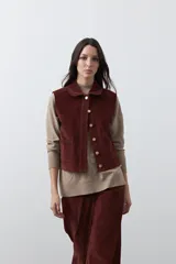 Chaleco de pana color marrón oscuro, con cuello camisero, cierre frontal de botones metálicos dorados y dos bolsillos frontales con detalle de bordado geométrico.