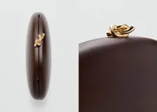 Bolso clutch rígido de madera color marrón, de forma ovalada, con cierre superior metálico dorado con detalle de nudo y forro interior de poliéster.