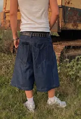 Bermuda de jean estilo raw denim, confeccionada en algodón de 12 onzas. Presenta un parche de cuero grabado con diseño barroco en la parte posterior y costuras reforzadas en contraste.