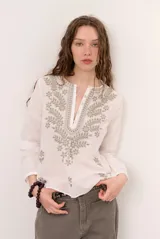 Blusa blanca de manga larga con cuello en V y cierre frontal de botones. Presenta delicados bordados en tonos grises y verdes alrededor del escote y en el pecho.