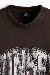 Remera de algodón color marrón con efecto lavado, cuello redondo y estampado frontal con la palabra 'SUNSET' en estilo universitario desgastado.