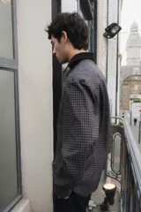 Camisa de corte boxy con estampado de cuadros pequeños en tonos oscuros. Presenta cuello camisero en color negro, cierre frontal con botones, canesú en la parte superior y un bolsillo en el pecho con etiqueta de la marca Kapsul Studio.
