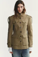 Chaqueta de estilo militar en color beige, con cuello subido, cierre frontal de botones, bolsillos de plastrón con solapa en el delantero y detalle de solapas en el pecho. Presenta mangas largas con hombreras y trabillas decorativas.