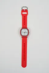 Reloj con correa de silicona roja, hebilla metálica plateada y caja cuadrada de borde redondeado. La esfera es blanca con números negros y tres subesferas.