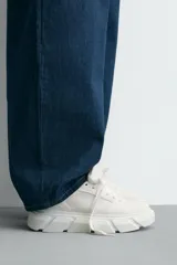 Zapatilla urbana de color blanco con diseño de suela gruesa con volumen y textura irregular. Presenta un corte superior con paneles combinados y cierre mediante cordones al tono.