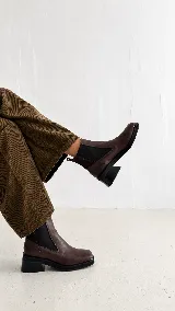 Bota tipo chelsea de cuero color marrón, con punta cuadrada y detalle metálico en la puntera. Cuenta con paneles elásticos laterales y taco cuadrado de altura media.