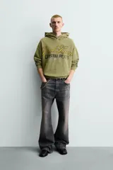 Sudadera con capucha de corte relajado en color verde oliva con efecto lavado. Presenta un bolsillo frontal tipo canguro y un estampado gráfico en el pecho con letras en tonos amarillos y negros. Cuenta con puños y bajo acanalados.