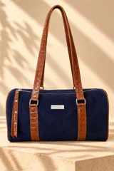 Cartera tipo baulito de cuero color marrón tostado, con doble asa larga de hombro, cierre superior y detalle de placa metálica con el logo de la marca en el centro.