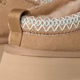 Zuecos UGG modelo Tazz de gamuza color beige, con plataforma alta y detalle de costura trenzada en el borde superior. Presenta el logo de la marca grabado en el lateral.