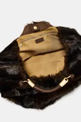 Maxi bolso de mano con acabado de pelo sintético en color marrón oscuro. Presenta un diseño estructurado con doble asa de mano reforzada y detalles metálicos dorados en las uniones. Cuenta con cierre mediante botón imantado y forro interior con bolsillo con cremallera.