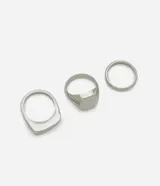 Kit de tres anillos de metal plateado: uno de sello con superficie ovalada, uno con superficie cuadrada y uno de banda simple y fina.