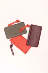 Monedero rectangular de cuero vacuno con textura granulada en color marrón topo. Cuenta con cierre superior metálico y un tirador decorativo de cuero en color naranja con un nudo. Presenta un pequeño logo grabado en el centro.