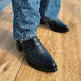 Zueco de cuero vacuno color negro, con diseño de tira trenzada en el empeine y cierre con hebilla metálica en el talón. Presenta punta fina y taco cuadrado de madera con acabado en cuero.