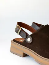 Zueco de cuero gamuzado color chocolate, con diseño de punta cerrada y tira ajustable con hebilla metálica en el talón. Presenta una suela de plataforma dentada en tono marrón claro.