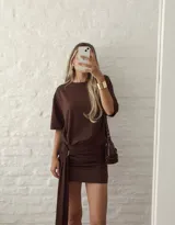 Vestido corto color marrón, con diseño de cuello redondo y mangas cortas holgadas. Presenta un detalle de fruncido en la cintura con una argolla metálica y una tira larga colgante que aporta movimiento.