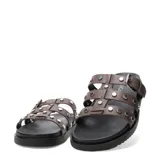 Sandalias planas Miss Carol modelo Nevada, color marrón, con tres tiras en el empeine decoradas con tachas metálicas plateadas. Una de las tiras cuenta con una hebilla de ajuste lateral. Poseen suela y plataforma baja de color negro.