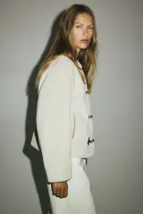 Campera de tejido suave efecto peluche en color crudo, con cuello redondo, hombros caídos y cierre frontal mediante alamares con detalles en contraste negro. Cuenta con bolsillos laterales ribeteados y forro interior.