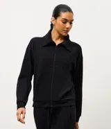 Campera deportiva negra con cierre frontal, cuello tipo polo y paneles acanalados en el pecho. Presenta mangas largas con puños elásticos y un diseño moderno y funcional.