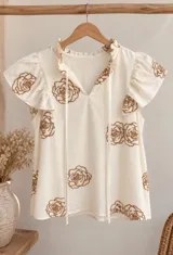 Blusa blanca de lino sin mangas, con escote en V y cuello con volados y tiras colgantes. Presenta bordados en hilo marrón con diseño de rosas distribuidos por toda la prenda y mangas con volados suaves.