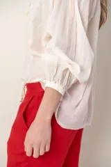 Camisa color crema de corte holgado, con cuello mao, cierre frontal con botones y volantes, mangas largas con puños elásticos y bordados en hombros y puños.