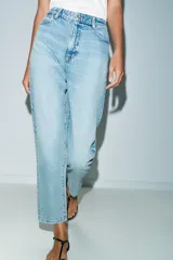 Jeans de denim azul claro lavado, tiro alto, corte Mom Fit con pierna recta y largo tobillero. Presenta cinco bolsillos y cierre frontal con cremallera y botón.