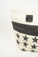 Bolso tipo tote de material textil con textura rústica, color beige con estampado de estrellas negras y una franja horizontal de glitter negro. Cuenta con asas largas de color claro y detalles de parches cuadrados negros en la unión de las asas.