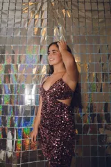 Vestido corto de fiesta color plateado con acabado brillante. Presenta un diseño de dos piezas que simula un conjunto de top y pollera, con aberturas laterales en la cintura, escote en V y tirantes finos que se cruzan en la espalda.