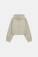 Sudadera tipo bomber de tejido suave, color beige, con cuello alto ajustable mediante cordones, cierre frontal con cremallera, mangas largas con puños elásticos y bolsillos laterales de vivo.