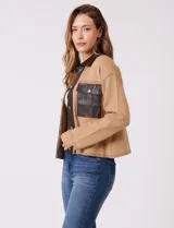 Chaqueta de corte boxy confeccionada en tejido de punto color crema, con detalles en cuero sintético negro en cuello, solapas, cartera central y bolsillos frontales. Presenta cierre frontal con botones metálicos plateados y costuras decorativas visibles.