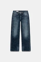 Jeans de tiro bajo y corte wide leg, confeccionados en denim azul con efecto lavado. Presentan detalle de tachas metálicas a lo largo de las costuras laterales y diseño clásico de cinco bolsillos.