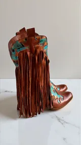 Botas tejanas de cuero color marrón terracota, con detalles pintados a mano en tonos turquesa y negro, diseño con flecos largos y puntera perfilada.