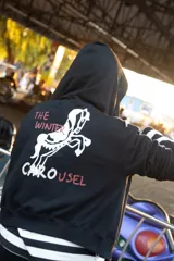 Campera con capucha de color crema, con diseño estampado en la espalda que incluye un caballo y la frase The Winter Carousel. Presenta bandas laterales con inscripciones en las mangas y un borde inferior con franjas horizontales en tonos bordó y celeste.