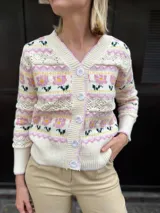 Cárdigan de punto con escote en V y cierre frontal de botones. Presenta un diseño tejido con motivos florales en tonos rosa, amarillo y verde sobre una base color crema, con detalles de calados horizontales.