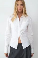Camisa de manga larga en tejido popelín color blanco, con cuello clásico, cierre frontal de cremallera metálica y bolsillos frontales tipo parche. Presenta un diseño entallado con abertura en el bajo.